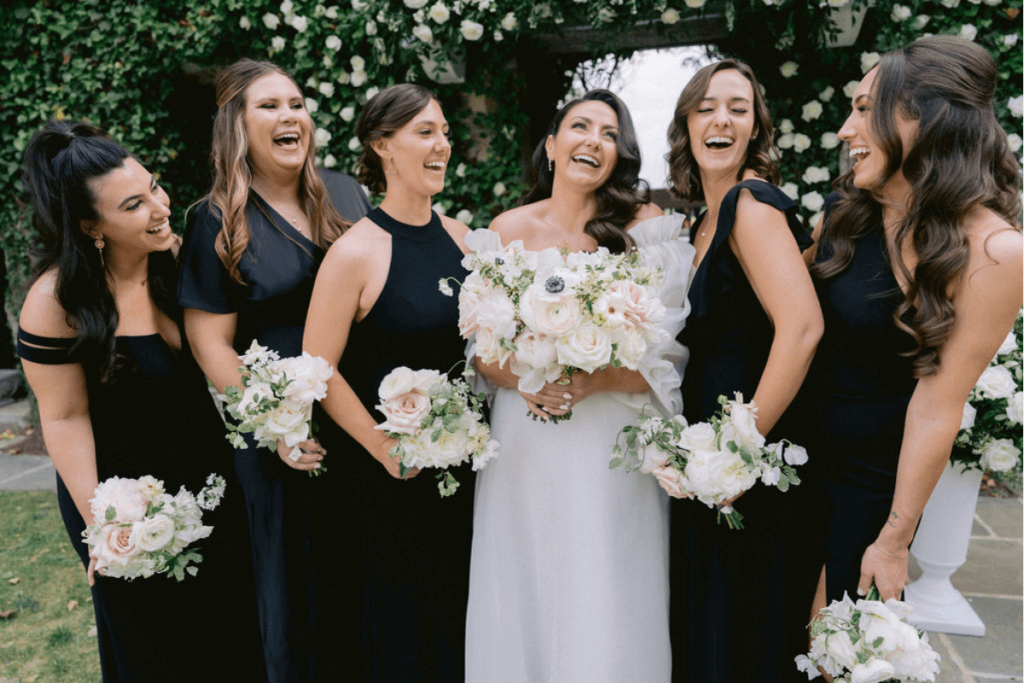 Image of a bride and her five bridesmaids wearing form black dresses laughing - this is an example of an energetic shot from a wedding photographer shot list
